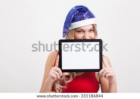 The beautiful blonde with big breasts standing on a white background and holds a a tablet in blue New Year's cap and smiling at the camera, selective focus on the tablet, picture with depth of field