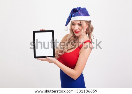 The beautiful blonde with big breasts standing on a white background and holds a a tablet in blue New Year's cap and smiling at the camera, selective focus on the tablet, picture with depth of field