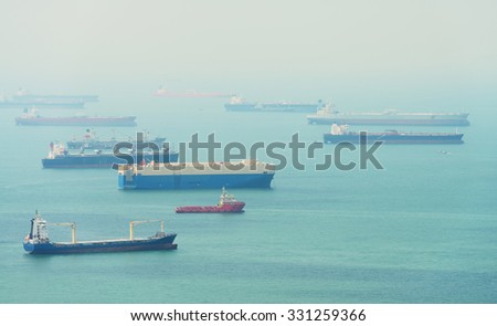 Many enormous, commercial cargo vessels of various types and sizes, moored in a harbor on a hazy day.