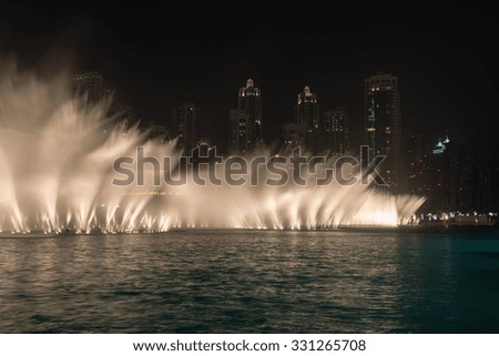 Dramatically lit fountains throwing white curtains of water into the air along the tropical beachfront of a major metropolitan city at night.
