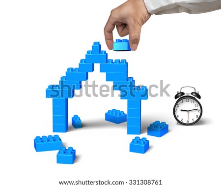 Human hand holding a blue block to complete house shape with alarm clock, isolated on white background.
