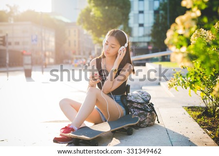 Young handsome asiatic long hair woman skater sitting on a small wall in the city backlight, listening music with headphones, holding a smartphone, looking down the screen - technology, relax concept