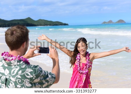 Man taking pictures with smart phone of woman on beach on Hawaii. Young couple having fun living happy lifestyle on Hawaiian beach holiday vacation travel. Man using smartphone for photo.