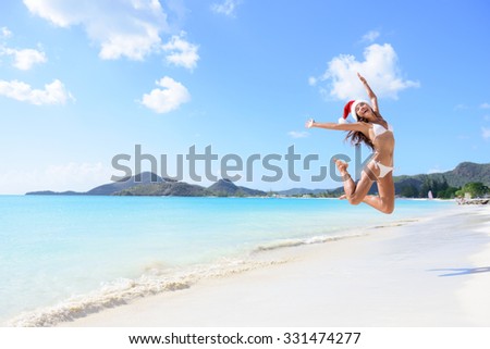 Happy Christmas vacation - girl jumping of joy and surprise on perfect white sand beach for winter holidays. Young woman wearing santa hat arms raised of happiness during vacations.