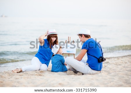 Woman, man and child in blue and white dress sit on the beach and draw on the easel near the surf