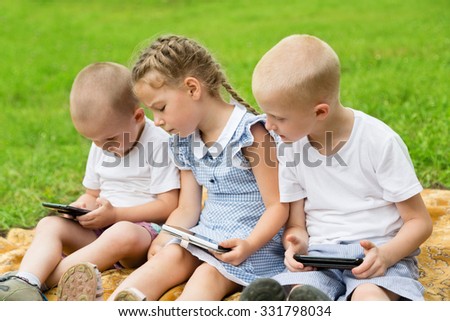 Brothers and sister using smartphones sitting on the lawn in park. Summer park. 