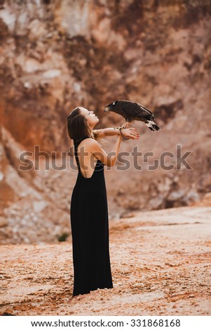 Amazing lifestyle picture of hippie woman holding eagle on her hand.looking away from camera.wearing stylish colorful boho style outfit .young girl in black dress standing in the mountains,expedition