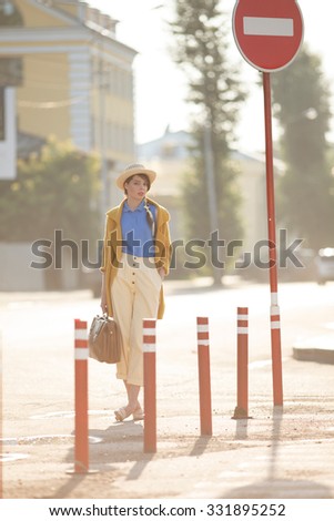 Young happy funny (vintage) dressed woman with retro suitcase stands on the street near red small columns and road sign. Picture ideal for illustating woman magazines.
