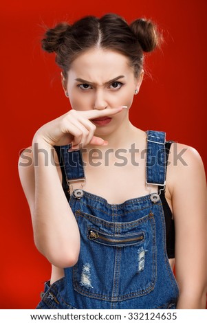 Angry young woman, with top knot hairdo, wearing on denim jumpsuit, is posing with finger near her nose, on the red background, in studio, waist up