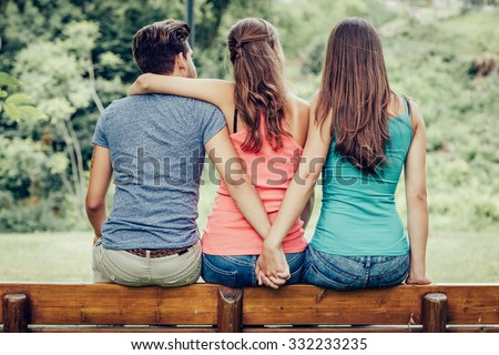 Love triangle, a girl is hugging a guy and he is holding hands with another girl, they are sitting together on a bench Royalty-Free Stock Photo #332233235
