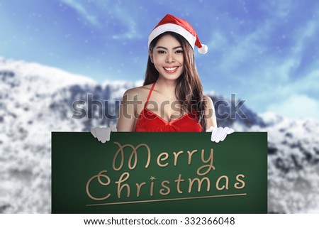 Young attractive asian woman wearing Santa Claus costume holding billboard with merry christmas writing