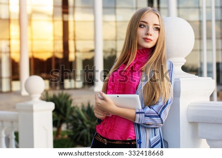 Young blonde businesswoman checking her tablet computer as she stands outdoors in the street in front of an office building windows