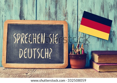 a chalkboard with the question sprechen sie deutsch? do you speak german? written in german, a pot with pencils, some books and the flag of Germany, on a wooden desk Royalty-Free Stock Photo #332508059