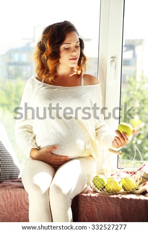 Pregnant woman sitting on window board in the room with basket of apples