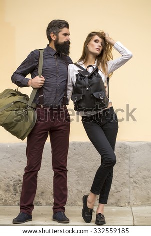 One beautiful stylish couple of young woman and senior man with long black beard standing embracing close to each other with travel bags outdoor on light orange wall background, vertical picture