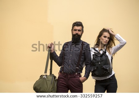 One beautiful stylish couple of young woman and senior man with long black beard standing embracing close to each other with travel bags outdoor on light orange wall background, horizontal picture