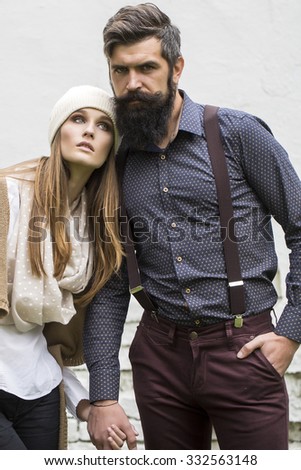 One beautiful stylish pensive couple of young woman and senior man with long black beard standing close to each other outdoor in autumn street on white brick wall background, vertical picture
