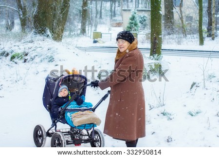 Great-grandmother walking with baby boy in pram during snowfall in winter. Happy family. Carefree childhood and generation.