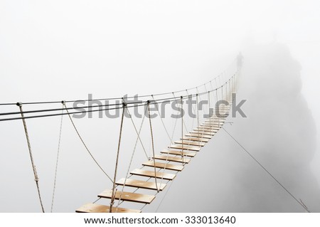 Fuzzy man walking on hanging bridge vanishing in fog. Focus on middle of bridge.  Royalty-Free Stock Photo #333013640