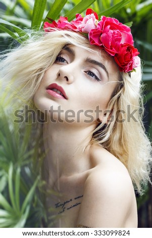 Close-up portrait of beautiful young woman in paradise, lost between palms. Red flowers in her hair, black tattoo on chest and gold tattoo on arm. White nails. Natural make up and relaxing feeling