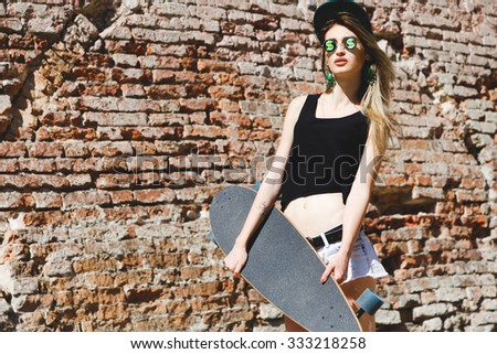 Young blond woman, with tattoo on her arm, wearing in shirt, shorts, cap and glasses, posing with skateboard, on the brick wall background, on the street, waist up