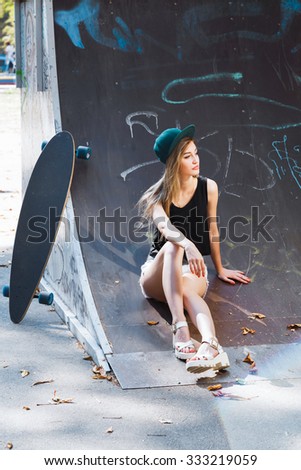 Attractive girl, wearing in shirt, sandals and shorts, sitting in the skatepark near her skateboard, full body