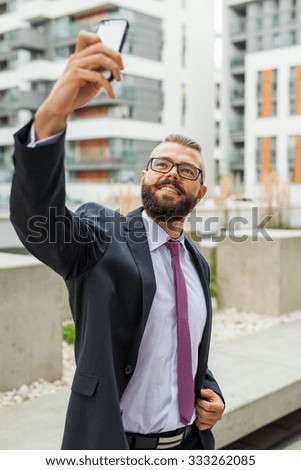 Young happy bearded businessman standing outside office building and taking selfie. Outdoor photo