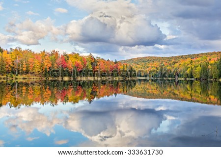 Vibrantly colorful fall foliage and white clouds in a blue sky are reflected on Lake Plumbago at Alberta in Upper Peninsula Michigan.