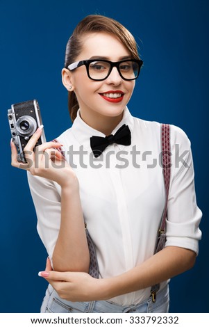 Smiling lovely girl, wearing in white shirt, glasses, brace, jeans and black bow, holding retro camera in her hand, on the blue background, in studio, waist up