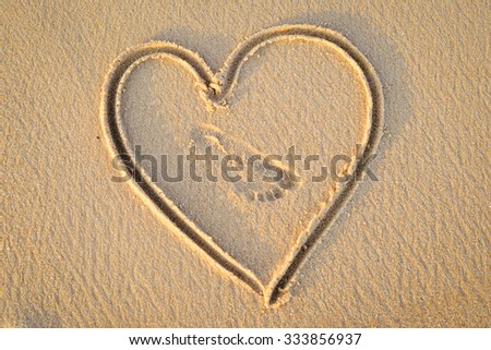 Picture of heart handwritten on golden sand. Sign of love with foot print in it on sunny outdoor background.  