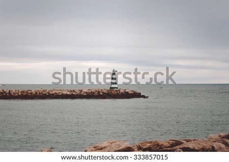 Picture of striped lighthouse on rocky pier in cold sea. Symbol of hope on sombre sky and grey water background.