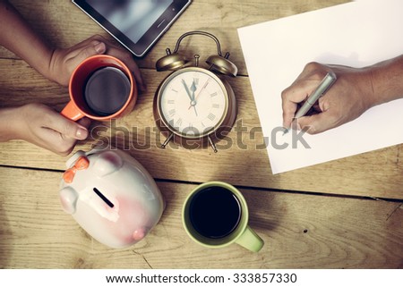 Picture of man's hand writing and woman's hands holding cup of coffee beside piggy-bank and alarm-clock. Top view of  family planning some purchasing on wooden plank table background.