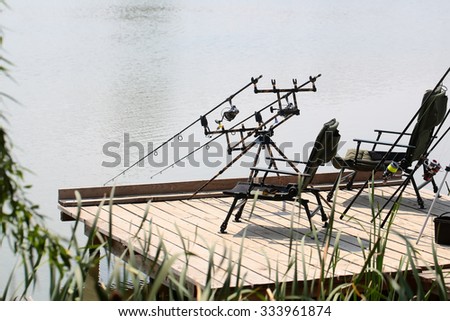 Black portable armchairs for rest and fishing pod with carbon carp rods standing on pier at lake with smooth surface of water in spring season recreation calm nature, horizontal picture 