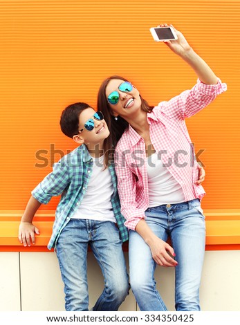 Happy mother and son teenager taking picture self portrait on smartphone in city, over colorful background, wearing a checkered shirt and sunglasses 