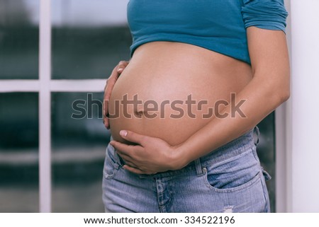 Nine months pregnant woman wearing jeans and a blue shirt stands in front of a window holding her hands on her bare belly.