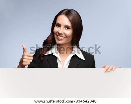 Portrait of happy smiling young businesswoman in black suit, showing blank signboard with blank copyspace area for slogan or text, over grey background, showing thumb up gesture
