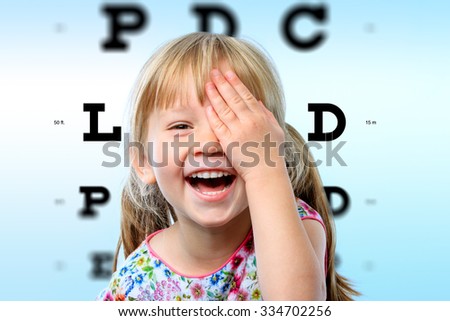 Close up face portrait of happy girl having fun at vision test.Conceptual image with girl closing one eye with hand and block letter eye chart in background.