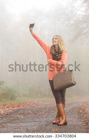 Happy fashion woman in fall autumn park taking selfie self photo picture. Pretty joyful young girl in sweater pullover with handbag photographing.