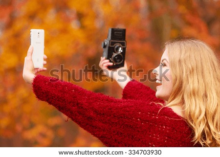 Pretty woman in fall forest park taking selfie self photo with old vintage camera and smartphone. Gorgeous young girl photographer. Autumn winter photography.