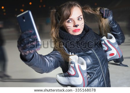 Healthy lifestyle and Halloween! Horror and magic holiday night. Pretty young woman with cat holiday makeup, make selfie on mobile phone. Girl holding ice skating boots