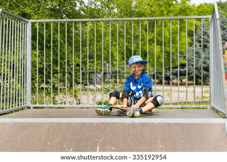 Smiling Young Confident Boy Sitting on Skateboard at Top of Ramp in Skate Park Surrounded by Green Trees
