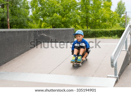 Front View of Young Boy Wearing Safety Pads Sitting on Skateboard and Riding Down Ramp in Skate Park Surrounded by Green Trees