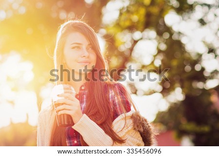 Happy teenage girl in autumn in park holding takeaway coffee. Beautiful young woman with coffee outdoors on sunny fall day. Vibrant colors, back light, shallow depth of field, medium retouch.