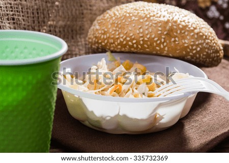Closeup still life disposable green cup white fork and cabbage and corn salad wheat bun topped with sesame seeds lying on brown brat on rustic background, horizontal picture 