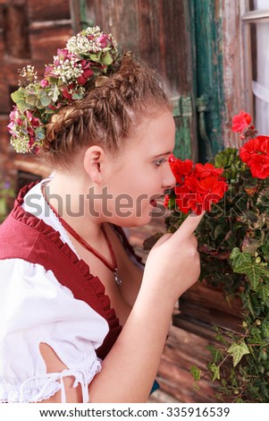 Portrait of a young chubby woman in red dirndl, you smelling a geranium in a flower boxes beneath a window / Portrait of a young woman in dirndl smell of a geranium