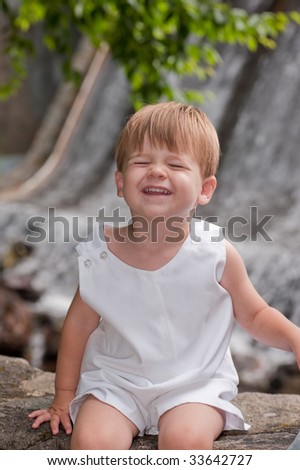 little boy sitting by a waterfall grinning at camera with eyes closed