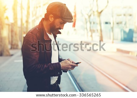 Half length portrait of bearded male with retro style using cell telephone while standing in urban setting, man dressed in stylish clothes chatting on smart phone during walking in cool spring day 