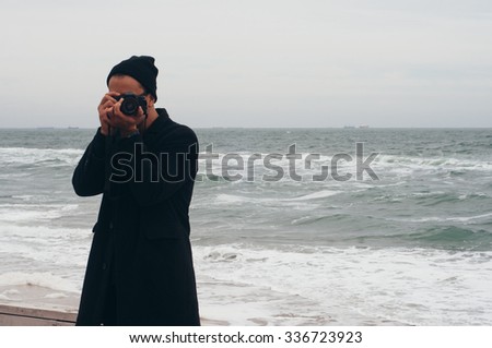 handsome hipster photographer traveler tourist guy wearing glasses and hat taking photo of hazy cold sea with his film camera 