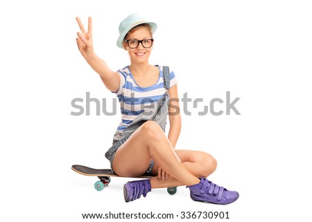 Studio shot of a hipster girl making a peace hand gesture seated on a skateboard isolated on white background