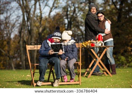 Cheerful little children sitting on the wooden chairs all together and looking through different family pictures on the tablet PC in the park.